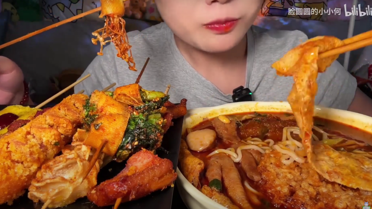 Screenshot from the YouTube channel of a mainland Chinese eating broadcaster, "Xiao He with a round face." The influencer is eating a large meal of noodle soup with numerous fried side dishes. 