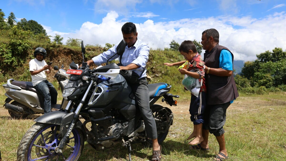 Man putting a young boy on a motorcycle. Another motorcyclist is in the background. Photo by Suman Nepali of Nepali Times, used with permission.